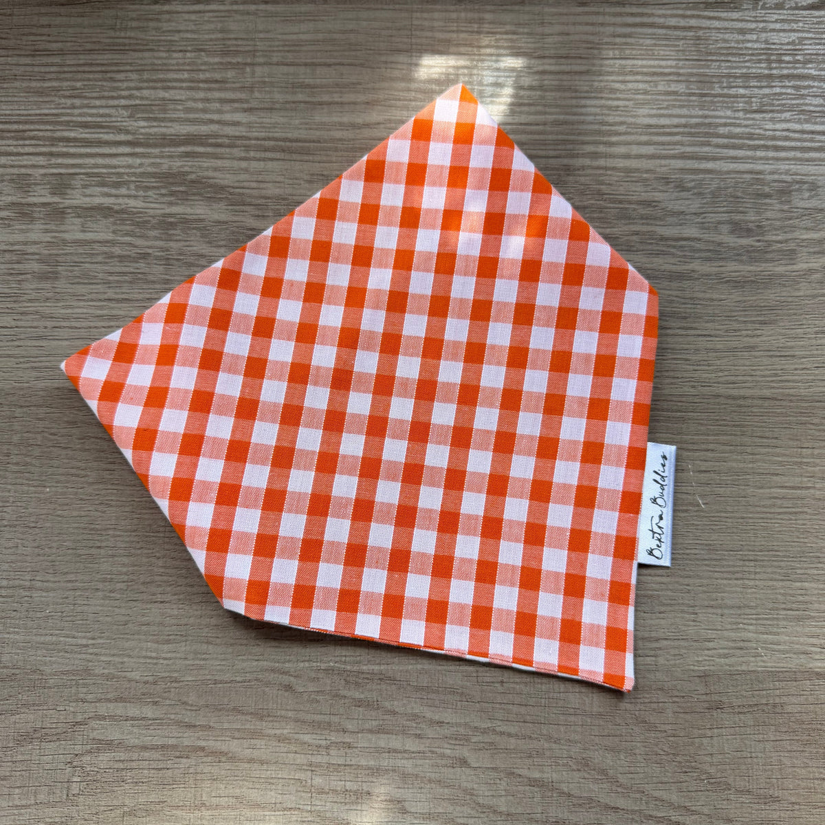 An orange and white gingham checkered bandana or pet bandana laid flat on a grey wood-grain surface, with a small white brand tag reading 'Bextra Buddies' visible on the right edge.