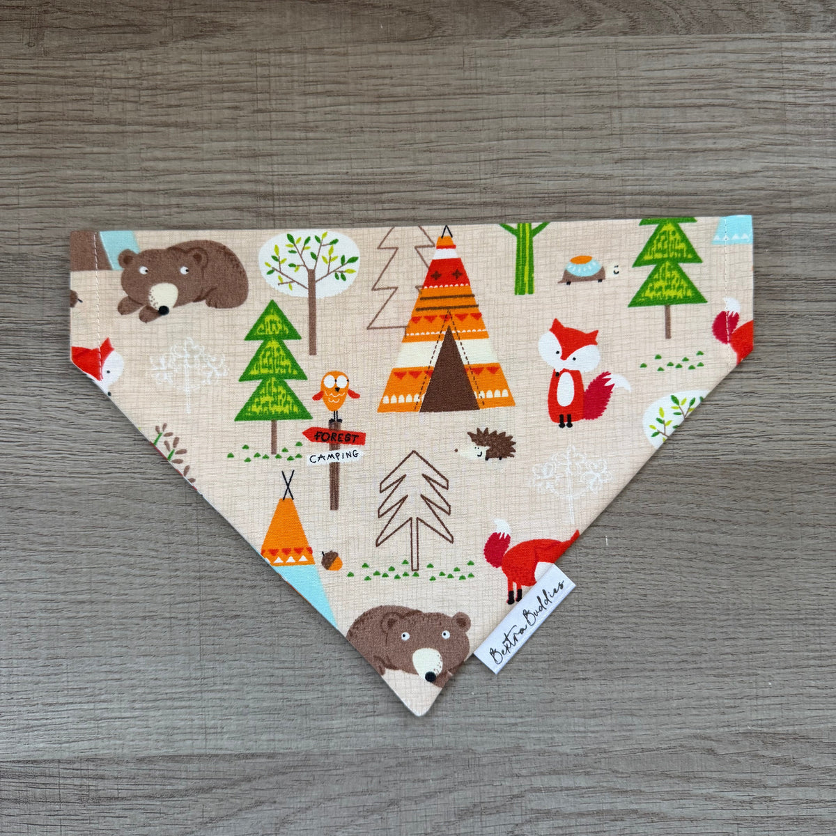 A triangular bandana-style pet bandana with a woodland-themed print, laid flat on a gray wood-textured surface. The print features brown bears, green trees, orange and brown teepees, and red/orange foxes on a light beige background.