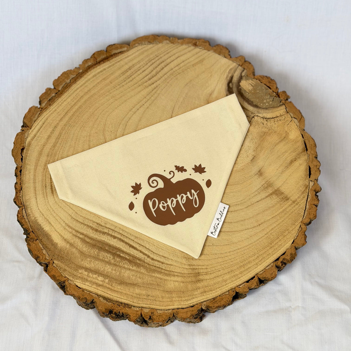 A cream-colored triangular bandana with a brown pumpkin graphic and the name 'Poppy' printed on it, styled on a rustic wood slice coaster against a white fabric background.