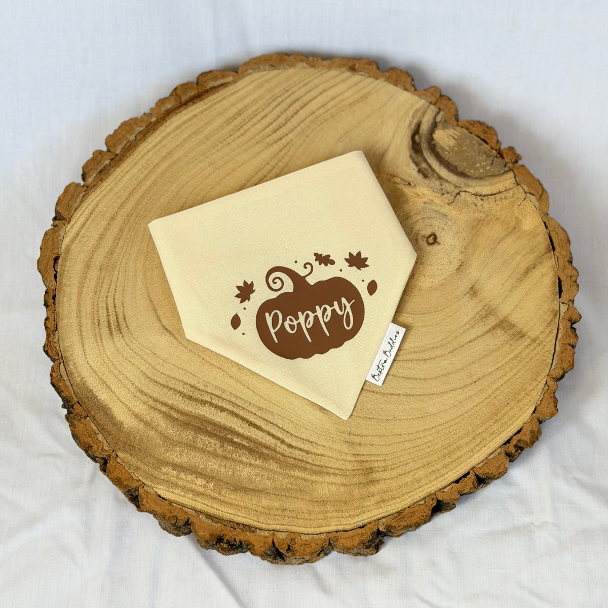 A cream-colored fabric bandana with a brown pumpkin and the word 'Poppy' printed on it, displayed on a round natural wood slice tray over a white fabric background.