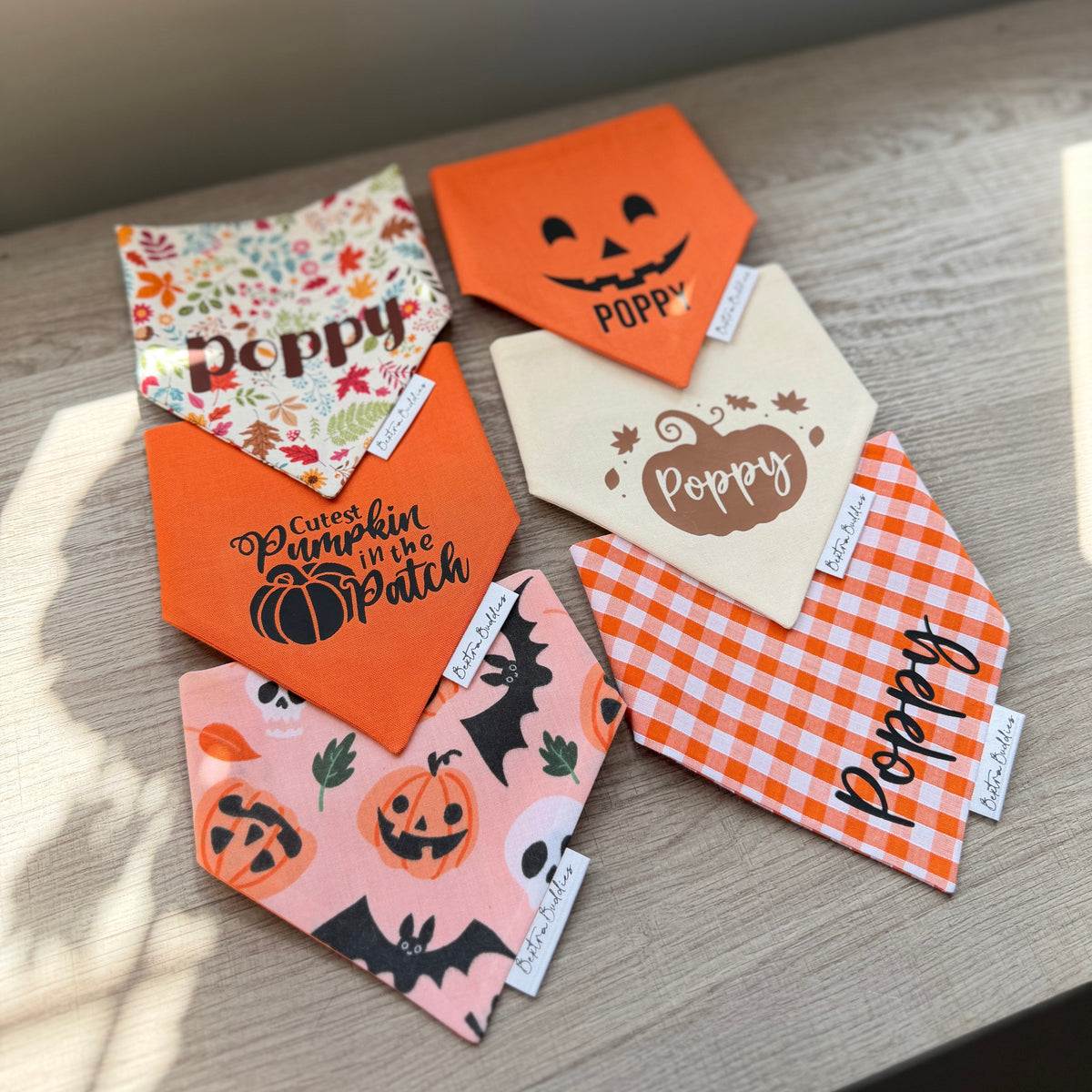 Personalized Halloween-themed bandana pet collars are arranged on a light wood surface. The collars feature various designs including orange pumpkin faces, a floral pattern with 'Poppy' written on it, 'Pumpkin in the Patch' with a pumpkin graphic, a pink pumpkin print with bats, and an orange-and-white gingham check pattern.