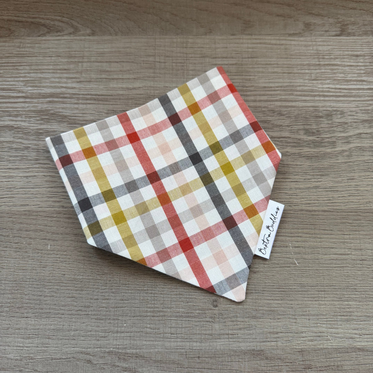 A small square fabric bandana or pet bandana with a multicolor plaid pattern, folded on a gray wood-grain surface. A small white tag with the brand name 'Bextra Buddies' is visible.
