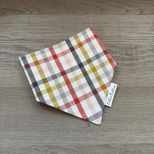 A small square fabric bandana or pet bandana with a multicolor plaid pattern, folded on a gray wood-grain surface. A small white tag with the brand name 'Bextra Buddies' is visible.