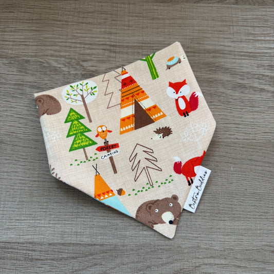 A triangular bandana-style pet bib with a woodland safari print, featuring cartoon bears, foxes, trees, and teepees on a beige background, displayed on a gray wood-textured surface.