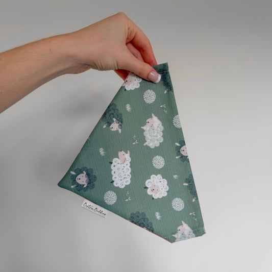Hand holding a green fabric dog bandana with floral sheep pattern on a plain background